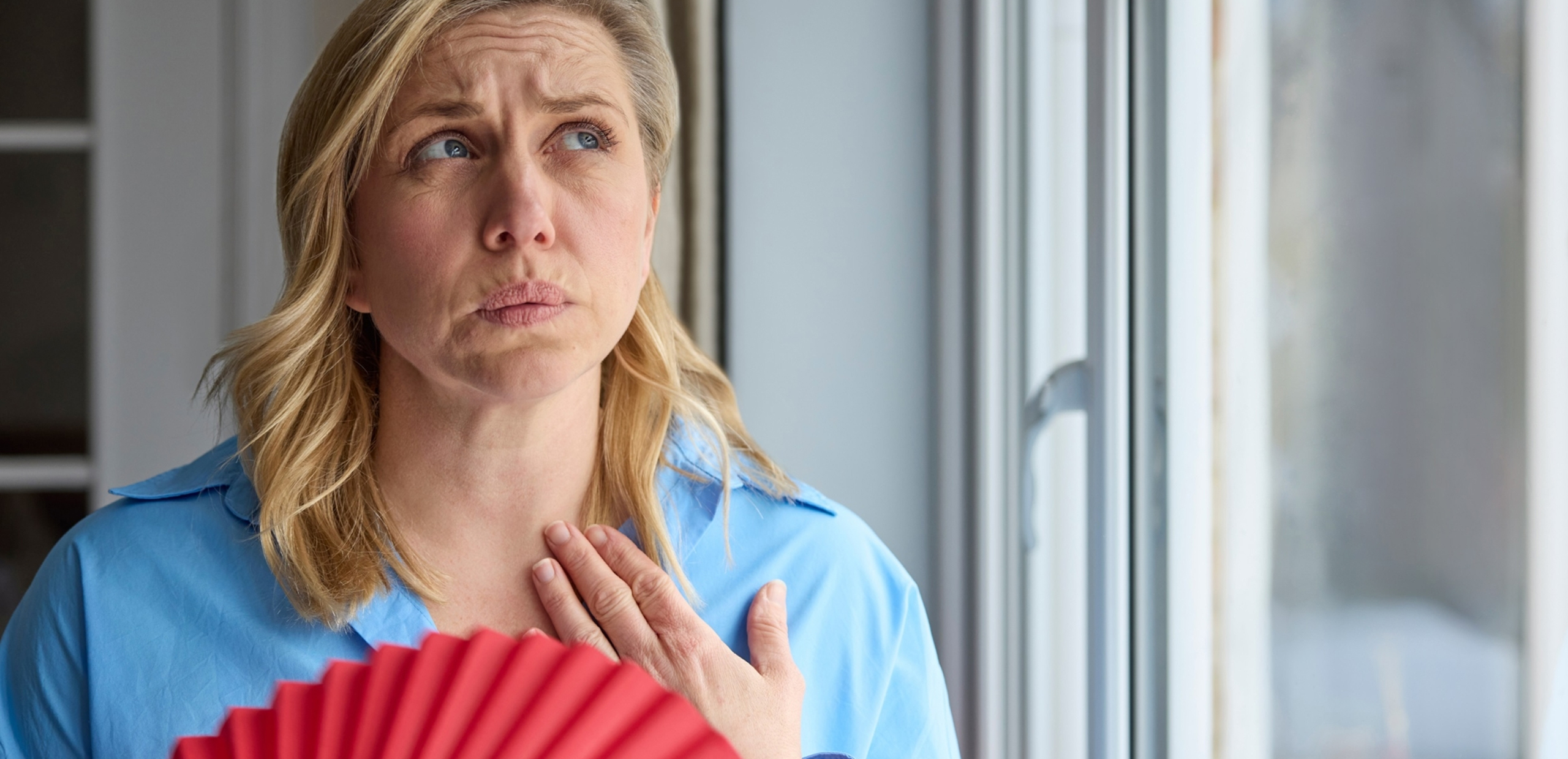 Middle-aged woman leaned on sofa resting in living room cools herself with orange colour hand fan touch forehead feels unwell suffers from unbearable hot summer weather at home without airconditioner